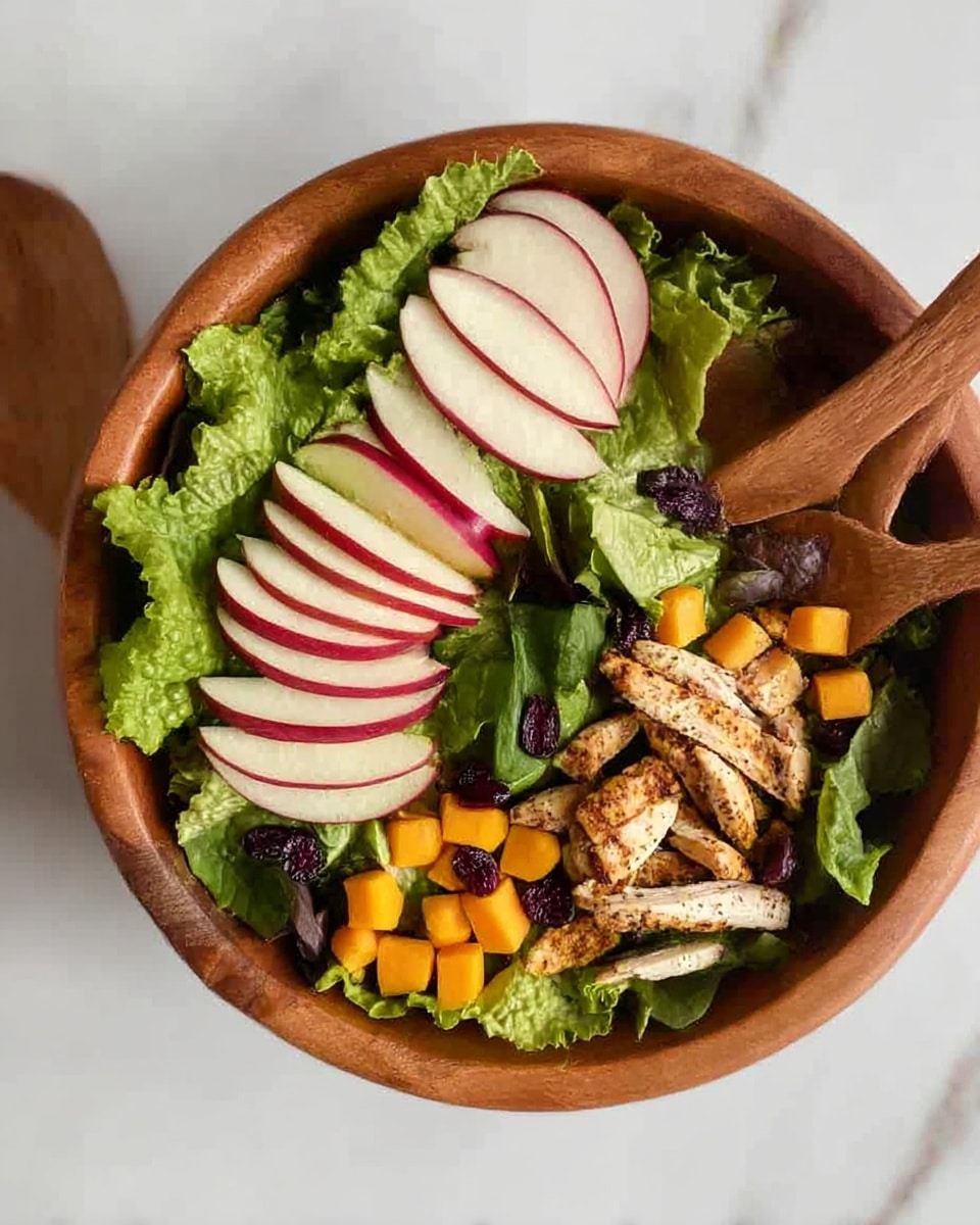A wooden bowl filled with a fresh salad sits on a white marbled surface, accompanied by wooden salad servers. The salad has four main parts: a base layer of green lettuce leaves with a ruffled texture, slices of light red apple with white flesh placed in two groups on opposite sides, small cubes of bright orange fruit or vegetable scattered around, and seasoned grilled chicken strips with a light brown color clustered near the bottom. Dark purple dried fruit or berry pieces are sprinkled lightly, adding contrast within the leafy greens. photo taken with an iphone --ar 4:5 --v 7