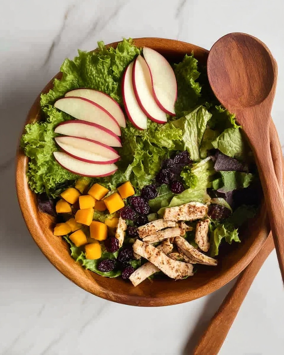 A wooden bowl filled with a fresh salad placed on a white marbled surface. The salad has a base layer of green leafy lettuce surrounding the edges, giving a ruffled texture. Inside the circle of lettuce, there are thin slices of red-skinned apple arranged in small fan shapes placed at three points around the bowl. Mixed green leaves with a variety of textures form the middle layer, interspersed with small chunks of bright yellow mango and dark little dried berries. At the center bottom of the bowl, there are strips of cooked light brown chicken with a slight crisp on the edges. There are also a few round pink slices peeking through the greens. Two wooden salad servers rest above the bowl on the white marbled surface. photo taken with an iphone --ar 4:5 --v 7