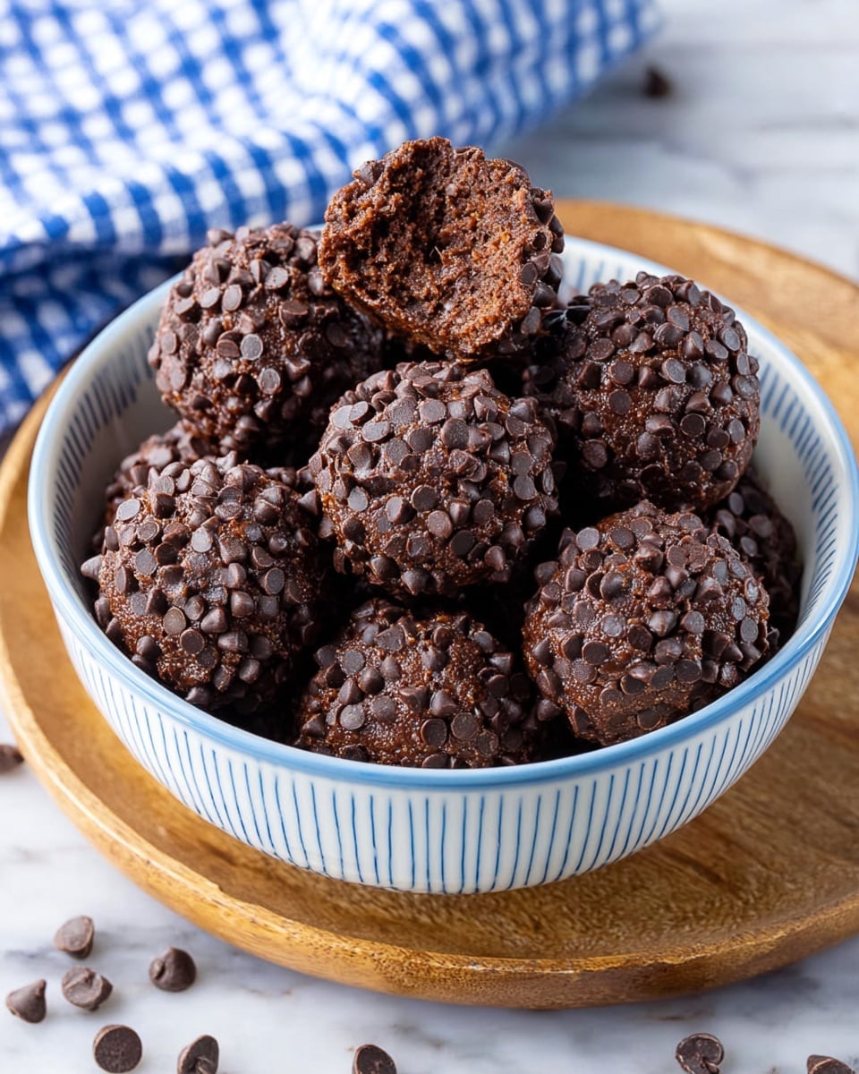 A white bowl with thin blue lines on the inside holds about eleven round chocolate treats, each covered in small, dark chocolate chips creating a bumpy texture on the surface. One treat has a bite taken out, showing a dense, dark brown inside. The bowl sits on a white marbled texture with some scattered chocolate chips around it, and a blue and white checkered cloth is partially visible in the background. photo taken with an iphone --ar 4:5 --v 7