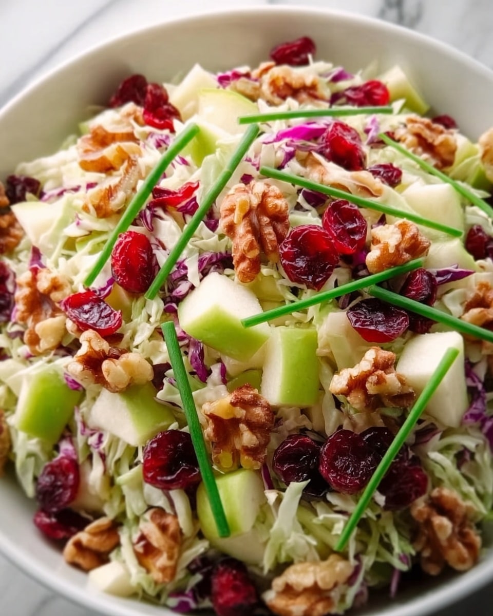 A close-up view of a colorful salad in a white bowl placed on a white marbled surface. The salad has several layers, starting with a base of shredded pale green cabbage and finely sliced red cabbage. On top, there are slices of light yellow apple, bright red dried cranberries, and chopped walnuts with a rough texture. Thin green chives are scattered across the salad, adding a fresh and vibrant touch. The overall look is fresh and mixed, with a variety of colors and textures visible. photo taken with an iphone --ar 4:5 --v 7