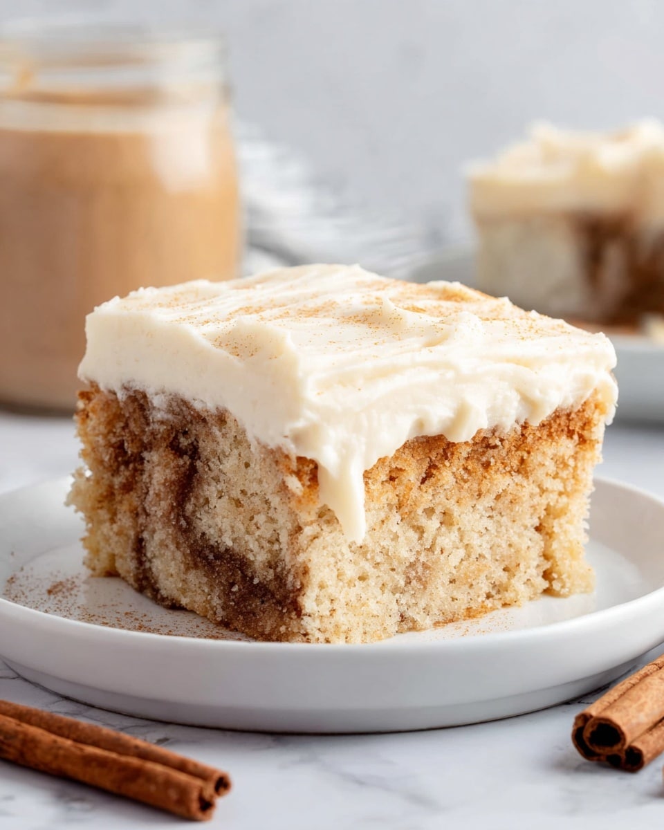 A thick slice of cake is shown on a white plate, sitting on a white marbled surface. The cake has two main layers: the bottom layer is a soft, light tan cake with visible swirls of darker cinnamon-colored filling mixed inside. On top, there is a thick, smooth layer of creamy, pale off-white frosting that spreads evenly across the surface. The texture of the cake looks moist and crumbly, while the frosting appears dense and rich. In the background, there is a cinnamon stick and a small blurred jar with brown contents, adding warmth to the scene. photo taken with an iphone --ar 4:5 --v 7