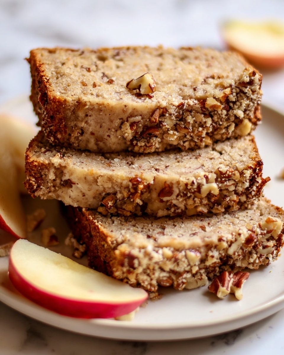 The image shows three slices of nut bread stacked on a white plate on a white marbled surface. The bottom slice is thick and light brown, with small nut pieces scattered inside. The middle slice is slightly thinner and has a crunchy top crust covered with finely chopped nuts, creating a rough texture. The top slice, also with a nut-covered crust, is placed at a slight angle. Around the slices, there are a few red apple wedges with a fresh and shiny skin. photo taken with an iphone --ar 4:5 --v 7