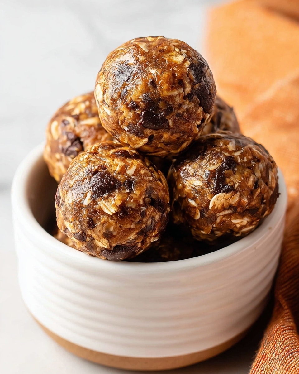 A close-up image of five round energy balls stacked inside a white ribbed bowl. Each ball has a textured surface showing visible oats and dark chocolate chunks embedded throughout, creating a mix of light brown and dark brown colors. The balls have a glossy, slightly sticky look, making them appear moist and fresh. The bowl is placed on a white marbled surface, with a hint of an orange-striped cloth on the right side adding a warm accent. Photo taken with an iphone --ar 4:5 --v 7