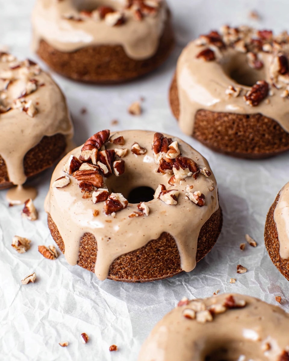 The image shows a close-up of several small, round donuts with a hole in the center, lying on a white marbled textured surface covered with crumpled white parchment paper. Each donut has two layers: a dark brown, textured base layer that looks soft and moist, and a thick, smooth, light beige glaze evenly coating the top, dripping slightly down the sides. The glaze is sprinkled with small pieces of chopped pecans, adding a rough texture and darker brown color contrast. The donuts are neatly arranged, filling most of the frame, with extra small nut bits scattered around. Photo taken with an iphone --ar 4:5 --v 7