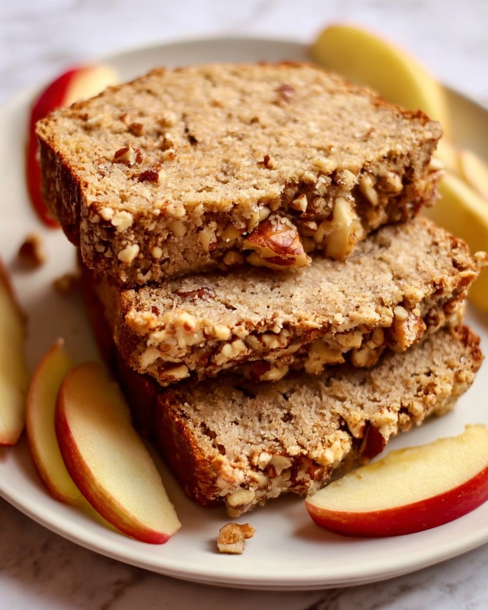 The image shows three slices of nut-studded bread stacked on a white plate with a white marbled texture underneath. The bread has a light tan color with visible pieces of nuts throughout the crumb and a slightly rough crust on top covered with crushed nuts, giving it a textured look. Around the bread slices, there are fresh apple wedges with smooth red and yellow skin, adding a warm contrast to the earthy tones of the bread. The lighting highlights the moist inside and crunchy outside of the bread. photo taken with an iphone --ar 4:5 --v 7