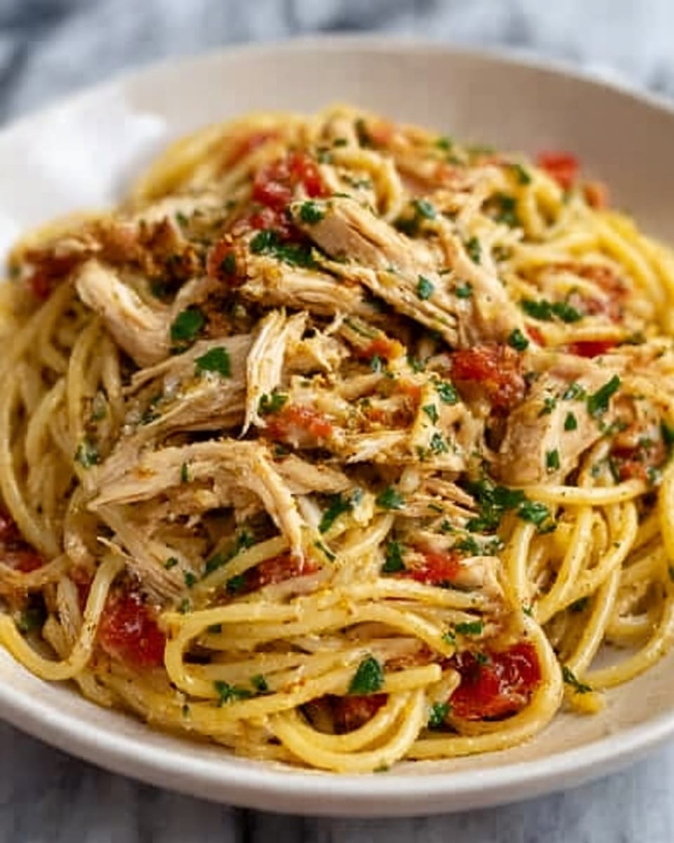The image shows a white plate filled with spaghetti mixed with shredded chicken pieces on top. The pasta is coated in a light sauce with visible bits of chopped red tomatoes and green herbs scattered throughout. The chicken pieces are golden and tender-looking, sitting on the pasta layers that have a smooth and slightly glossy texture. The dish looks fresh with a mix of warm yellow, red, and green colors, set against a white marbled surface. Photo taken with an iphone --ar 4:5 --v 7