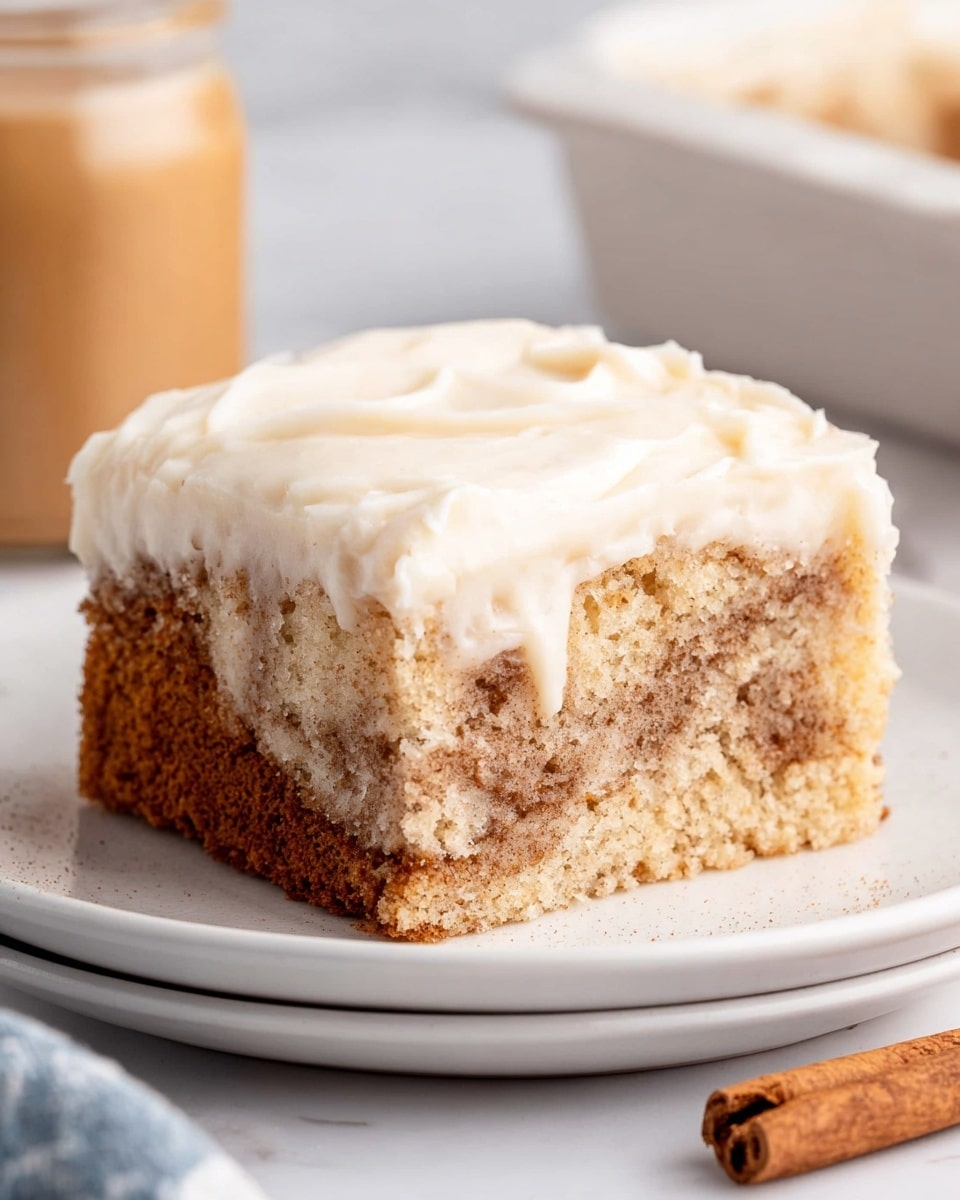 A close-up of a square piece of cinnamon cake on a white plate. The cake has two visible layers: the bottom layer is light brown with darker swirls of cinnamon and a crumbly texture, showing a mix of soft cake and spiced filling. The top layer is a thick, smooth, creamy white frosting, evenly spread and slightly fluffy with soft peaks. The plate rests on a white marbled surface with a cinnamon stick placed near the plate. A glass jar with brown contents is blurred in the background. photo taken with an iphone --ar 4:5 --v 7