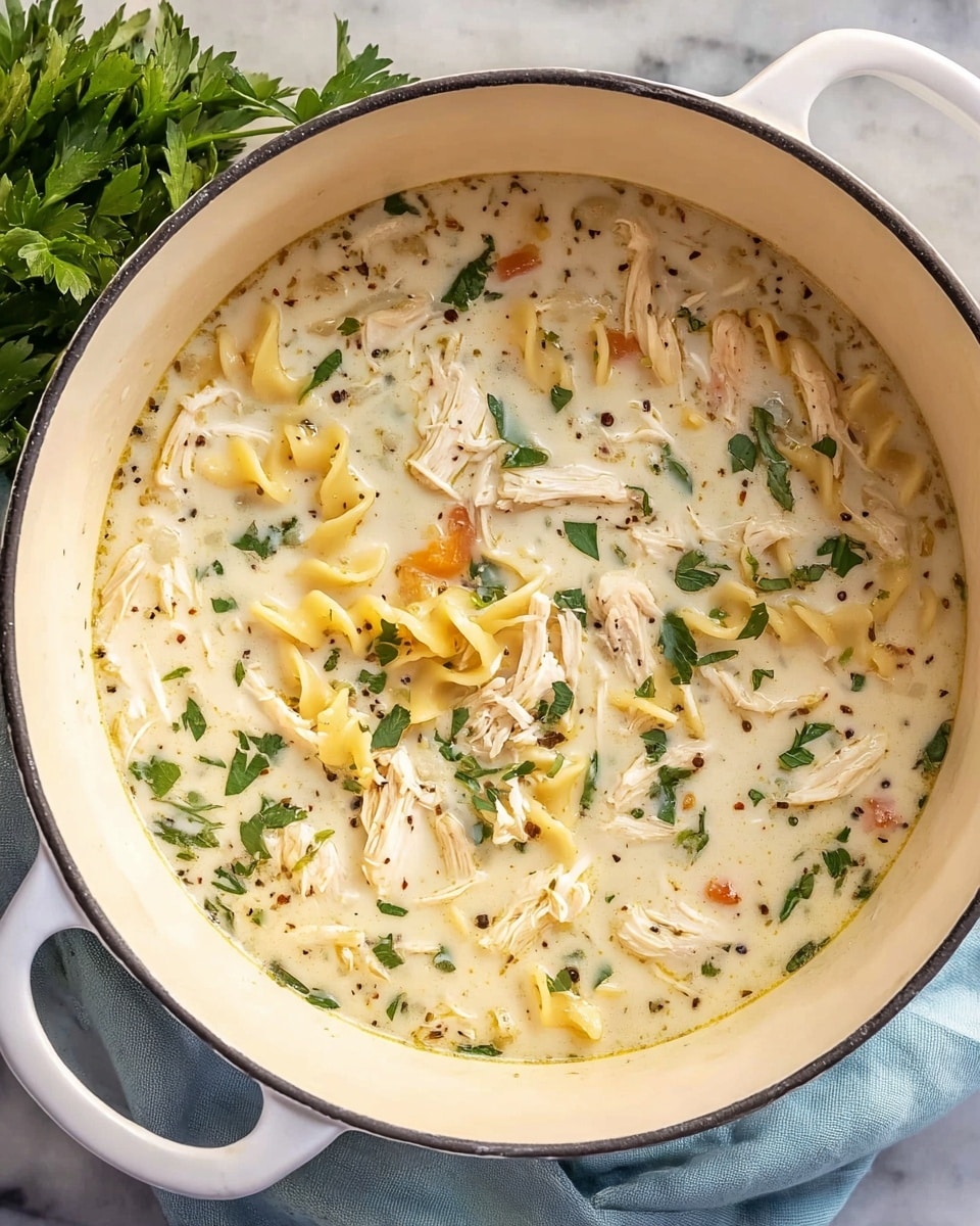 A white pot filled with creamy chicken soup showing three main layers inside: a light beige creamy broth base, scattered with shredded white chicken pieces, and small chunks of yellowish pasta. Fresh green parsley leaves float on top, adding a touch of bright green color, along with tiny dark specks of black pepper and herbs. The pot is placed on a white marbled surface with some fresh green parsley visible on the left edge and a light blue cloth underneath the pot. Photo taken with an iphone --ar 4:5 --v 7