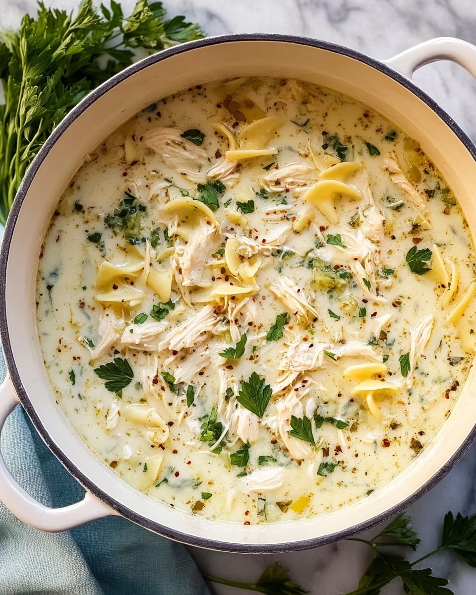 A creamy soup is shown inside a light blue pot with a white inside, placed on a white marbled surface with some green herbs on the side. The soup has multiple layers visible: the base is a pale creamy broth with a smooth texture, mixed with pieces of shredded white chicken evenly spread throughout. There are small pieces of yellow pasta scattered in the soup, along with green leafy herbs adding color contrast. Tiny black pepper specks and bits of brown spices are also sprinkled across the surface. The soup looks rich and comforting, filling the pot close to the top. photo taken with an iphone --ar 4:5 --v 7
