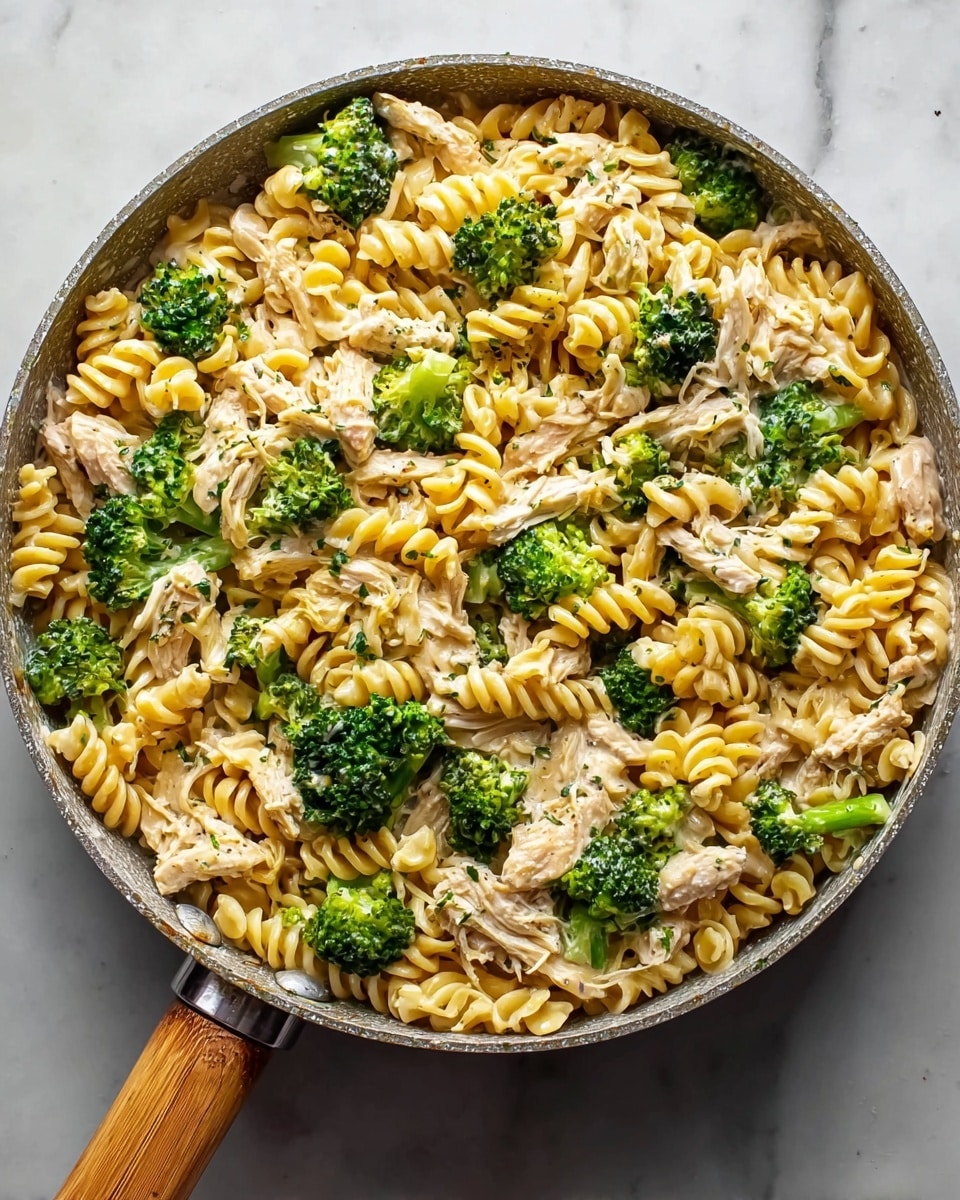 A large pan filled with a creamy pasta dish, showing three main layers mixed together: light yellow spiral rotini pasta, bright green broccoli florets, and shredded light brown chicken. The pasta spirals have a smooth texture and are well coated with a creamy sauce. The broccoli pieces add a fresh, rough texture and bright color throughout, contrasting with the soft shredded chicken that appears tender and juicy. These ingredients are evenly spread, making the dish look hearty and colorful. The pan has a slightly worn look with a dark rim and a wooden handle, sitting on a white marbled surface. photo taken with an iphone --ar 4:5 --v 7