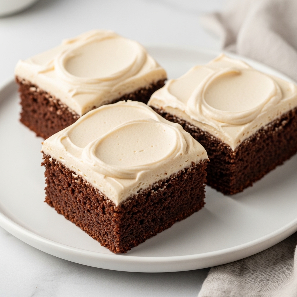 Three square pieces of dark brown chocolate cake with a thick, smooth layer of light beige frosting on top are placed closely on a round white plate. The dense cake base has a slightly crumbly texture while the creamy frosting is evenly spread with soft swirls and peaks. The plate rests on a white marbled surface with a soft cloth nearby. The overall look is simple and inviting. photo taken with an iphone --ar 4:5 --v 7