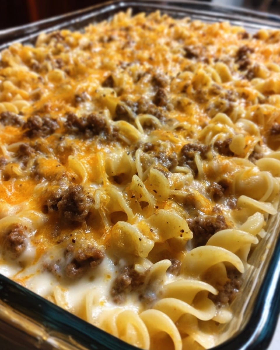 The image shows a close-up view of a baked pasta dish in a rectangular glass pan. The pasta is curly and covered with a golden-brown melted cheese layer sprinkled with small pieces of browned ground meat. Beneath the cheese and meat, the pasta appears creamy with a smooth texture. The white marbled surface serves as the background. Photo taken with an iphone --ar 4:5 --v 7