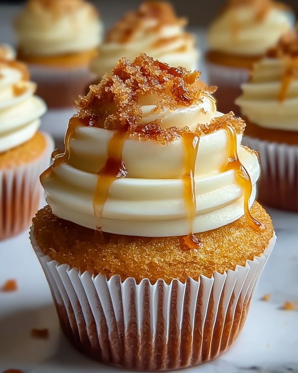 A close-up view of a single cupcake with a golden-brown textured base in a white cupcake liner. On top, there is a thick swirl of smooth off-white frosting that forms two layered rings, with the upper ring slightly smaller, giving a wave-like effect. The frosting is generously sprinkled with small, crunchy-looking caramelized brown sugar bits. A light drizzle of amber syrup runs down the frosting, adding shine and stickiness. In the blurred background, other cupcakes with similar frosting and toppings sit on a white marbled surface. photo taken with an iphone --ar 4:5 --v 7