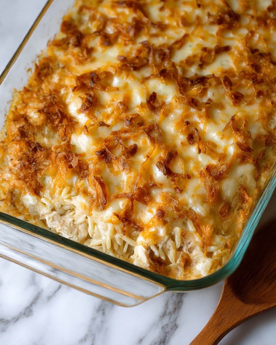 The image shows a baked casserole with at least two visible layers. The bottom layer consists of light golden pasta, possibly orzo, mixed with small pieces of creamy white chicken. On top, a thick layer of melted cheese spreads evenly, topped with crispy, browned fried onions scattered all over, giving a textured look with golden brown and darker toasted spots. The casserole is in a clear glass dish with white sides, placed on a white marbled surface. A wooden spoon is placed near the dish’s corner. Photo taken with an iphone --ar 4:5 --v 7