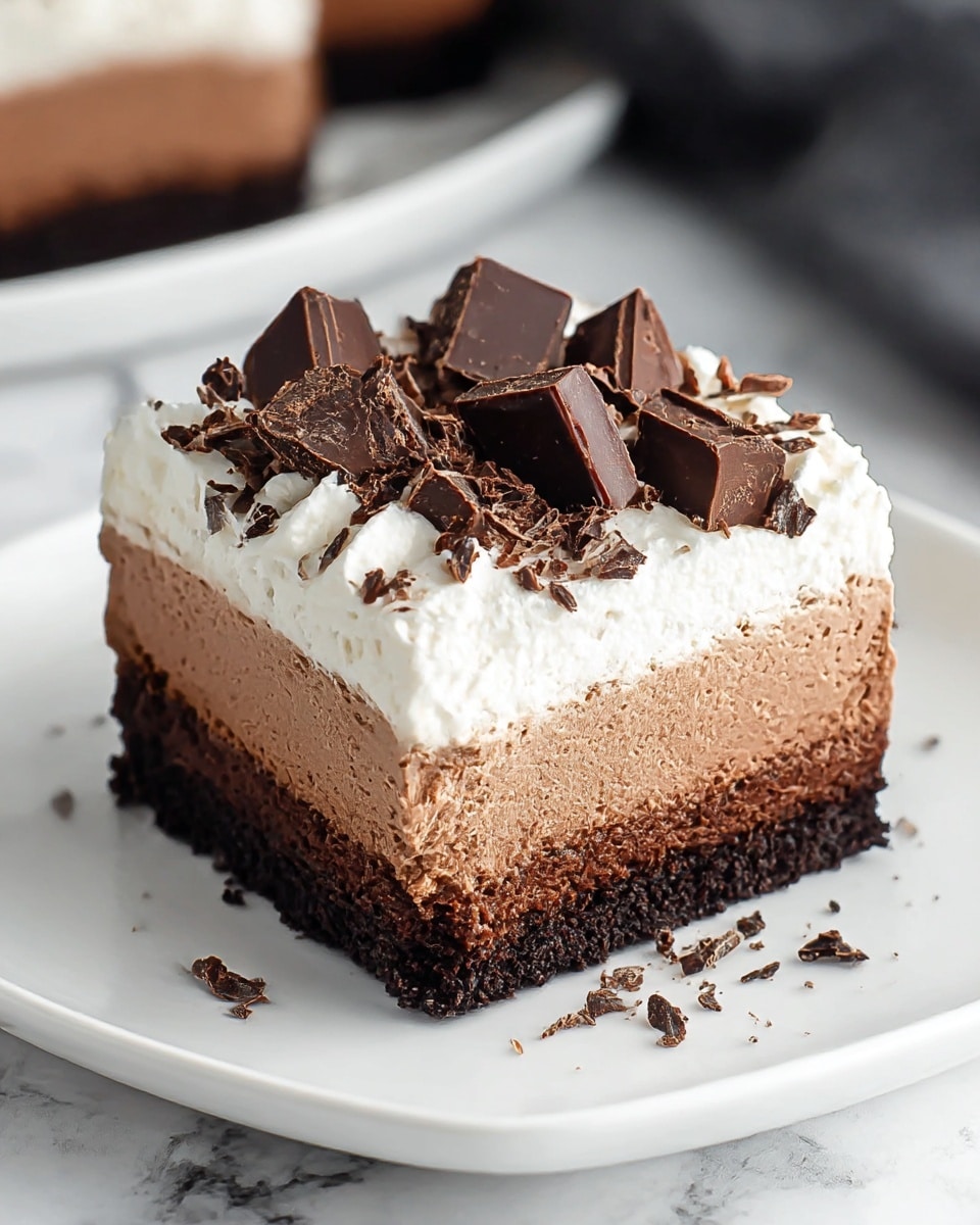 The dessert is a three-layered square chocolate mousse cake placed on a white plate, resting on a white marbled surface. The bottom layer is dark and crumbly like a rich chocolate cookie crust. The middle layer is a smooth, thick milk chocolate mousse with a soft texture and light brown color. The top layer is white whipped cream, soft and fluffy, spread evenly with slight peaks. On top of the whipped cream, there are various chunky pieces of dark chocolate arranged as decoration, along with small dark chocolate crumbs scattered lightly over the surface. photo taken with an iphone --ar 4:5 --v 7