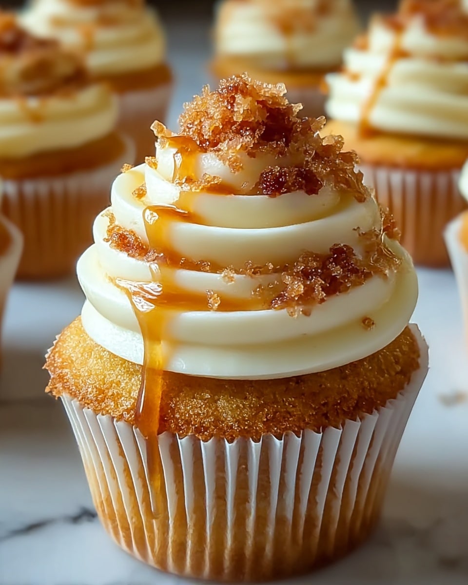 A close-up of a single cupcake shows three main layers: the bottom layer is a light golden-yellow moist cake wrapped in a white paper liner with vertical ridges, the middle layer is a swirl of smooth, creamy white frosting piped in multiple soft, thick waves, and the top layer features small, crunchy, caramelized brown sugar crystals scattered generously on the frosting with a light drizzle of golden syrup running down the sides. The background shows blurred cupcakes with similar layers on a white marbled surface. photo taken with an iphone --ar 4:5 --v 7