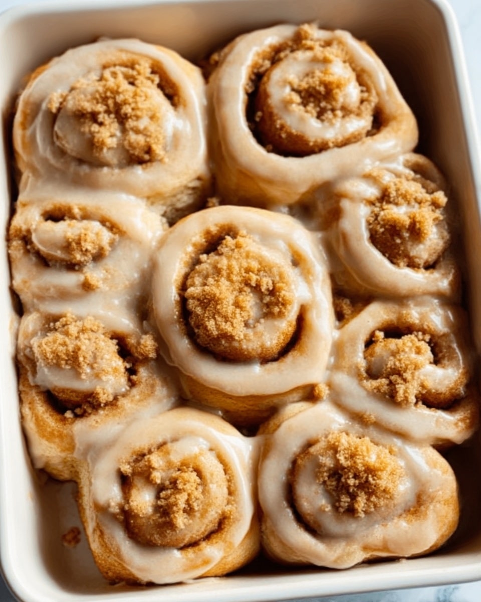 The image shows a white rectangular dish filled with nine cinnamon rolls arranged in three rows of three, each roll with smooth tan-colored glaze dripping slightly down the sides, creating a shiny texture. The cinnamon rolls have a light golden-brown baked dough color with swirled layers visible under the glaze. A sprinkle of crumbly light brown crumbs sits on top of each roll, adding texture. The white marbled surface underneath the dish provides a clean and bright background. Photo taken with an iphone --ar 4:5 --v 7