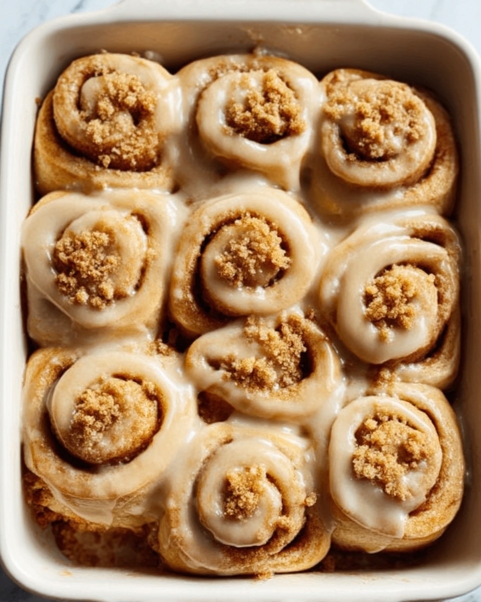 The image shows a close-up view of nine cinnamon rolls arranged neatly in a white rectangular baking dish, placed on a white marbled surface. Each cinnamon roll has a golden-brown color with swirled layers that create a spiral shape. The rolls are coated in a smooth, light tan icing that pools slightly into the crevices between the layers, giving them a shiny appearance. Crushed crumbs are sprinkled on top of each roll, adding texture and contrast against the smooth icing. The lighting emphasizes the softness and freshness of the rolls. Photo taken with an iphone --ar 4:5 --v 7